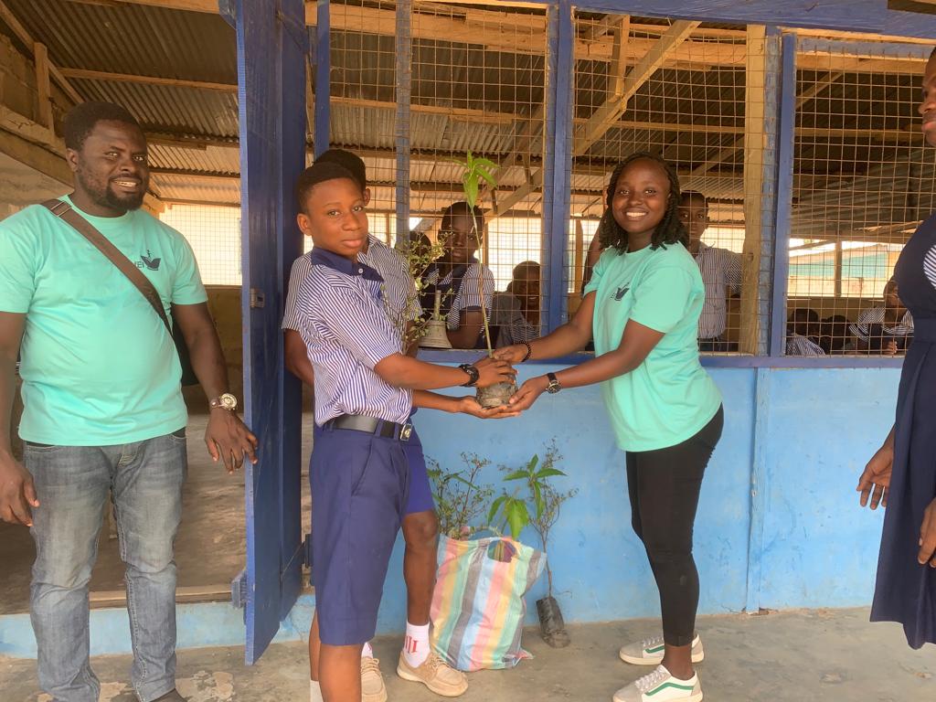 A facilitator in a teal T-shirt handing a tree seedling to a pupil in a classroom veranda, image 4 of 14